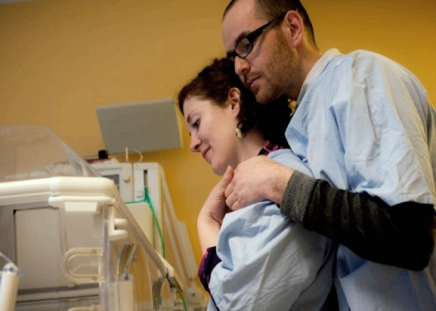 Bilde av et foreldrepar kledd i sykehusklær som ser ned på noen som er utenfor bildet, kanskje et barn i en sykehusseng