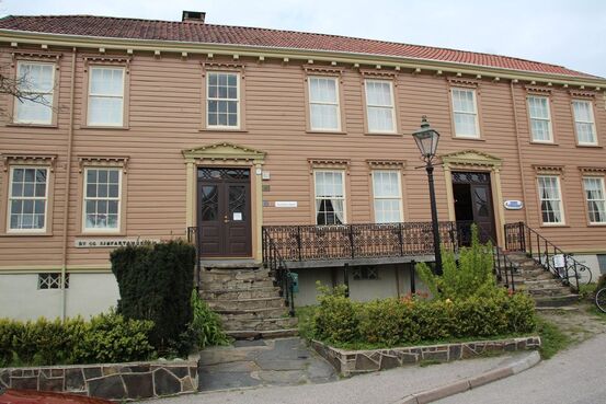 Lillesand by og sjøfartsmuseum lysebrunt bygg i to etasjer med hekker foran