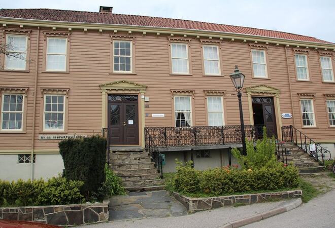 Lillesand by og sjøfartsmuseum lysebrunt bygg i to etasjer med hekker foran