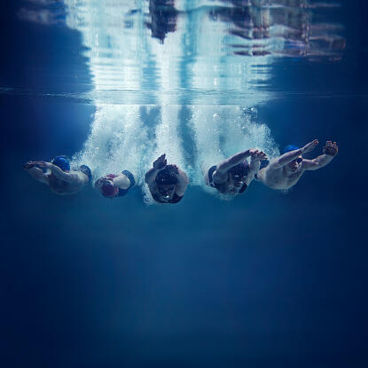 Five, young swimmers are jumping in direction of the camera. This is underwater shoot. There are 3 girls and 2 boys. They wear caps, swimming goggles and swimwears. The swimmers just entered the water with hands in front of themselves. They are looking do