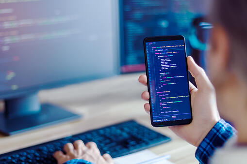 Closeup freelancer hand is holding smartphone with program code. Programmer developer is writing software on multiple computer screens at home office. Devices for working. Geek workplace concept. Closeup freelancer hand is holding smartphone with program code. Programmer developer is writing software on multiple computer screens at home office. Devices for working. Geek workplace concept.