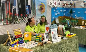 to jenter sit på stand med grøne tskjorter på med kommunelogo på, på bordet forran er det ulike bøker og ting knytt til barne- og ungdomarbeidarfaget