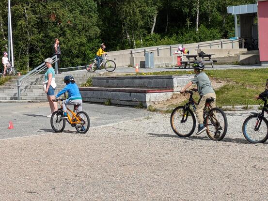 Fire elever i 1.-4. klasse sykler på skolegård, voksen passer på