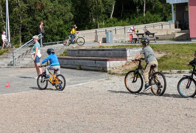 Fire elever i 1.-4. klasse sykler på skolegård, voksen passer på