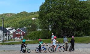 Fem elever fra 1. til 4. klass sykler på skolegård, en voksen person passer på