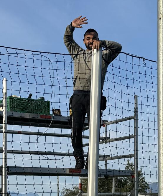 En mann med skjegg står på stillage og reparerer nett rund ballbingen