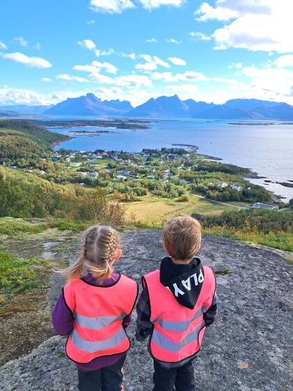 En gutt og en jente i ca 4-5 år i rosa refleksvester står på toppen av fjell og ser ned mot Skutvik