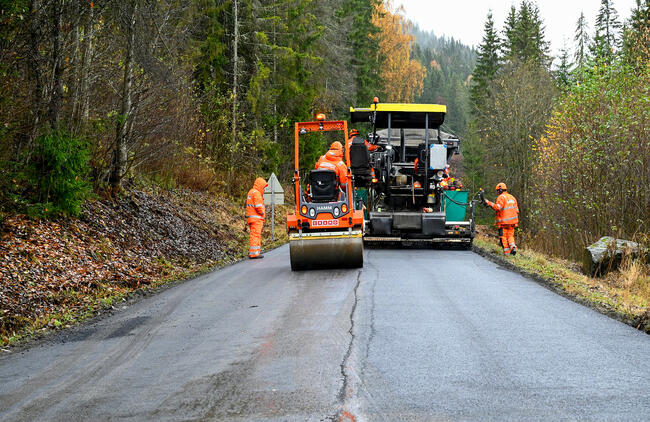 To maskiner asfalterer en vei i skogen, tre menn i orange klær passer på