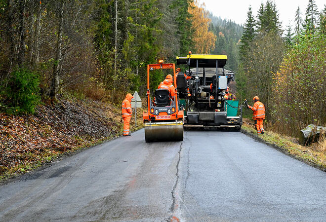 To maskiner asfalterer en vei i skogen, tre menn i orange klær passer på