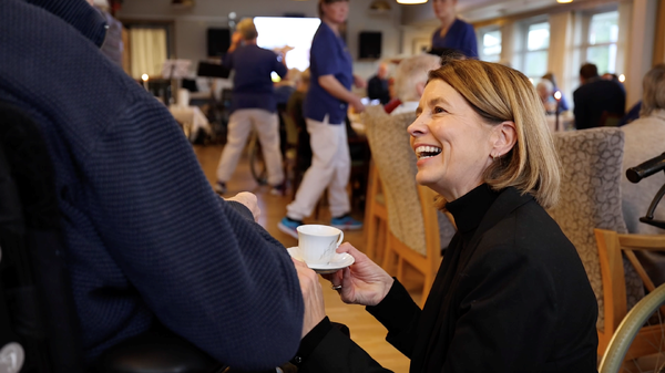 Ei smilande dame sit på huk ved sida av ein person som sit i stol, og gir ein kaffekopp.