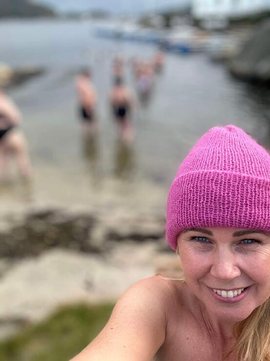 Person i forgrunnen med knallrosa lue og bare skuldre tar selfie. I bakgrunnen er der flere mennesker i badetøy som går ut i vannet.