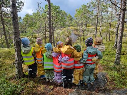 8 barnahage barn i regnklær og refleksvester står med ryggen mot oss i skogen