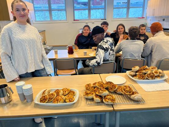 To jenter og fire guteer siter og spiser pizza på skolekjøkkenet. Ei jenta står ved bordet med pizza og smiler