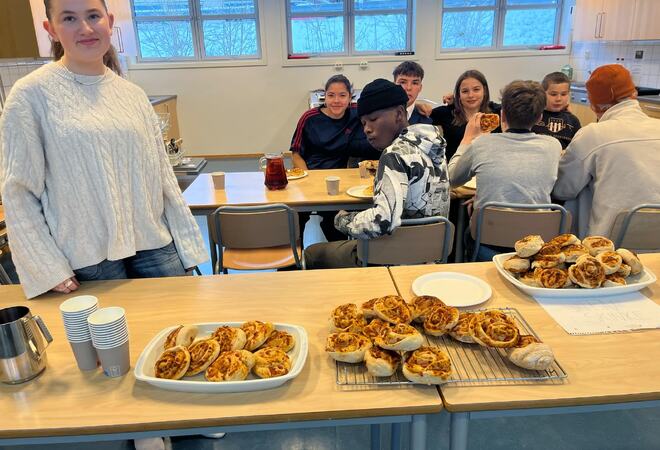 To jenter og fire guteer siter og spiser pizza på skolekjøkkenet. Ei jenta står ved bordet med pizza og smiler