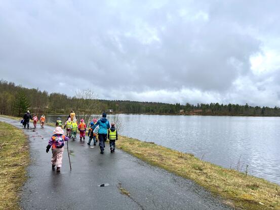 En gruppe barn og voksne i fargerikt regntøy går langs en våt sti ved en innsjø, omgitt av trær
