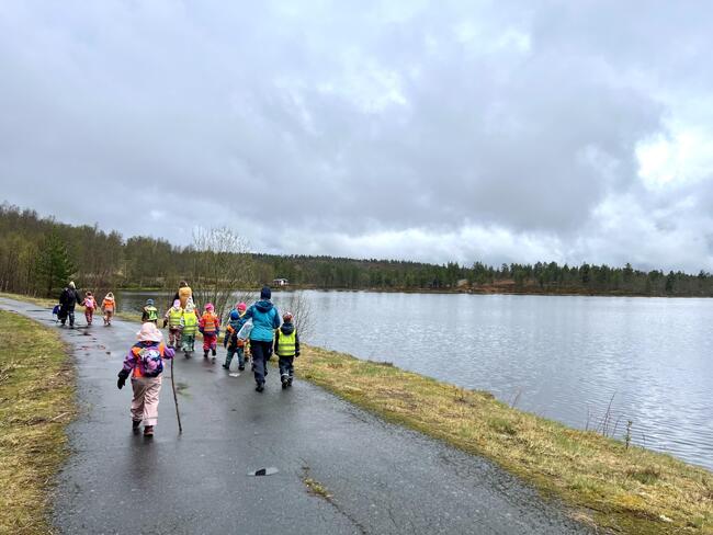 En gruppe barn og voksne i fargerikt regntøy går langs en våt sti ved en innsjø, omgitt av trær