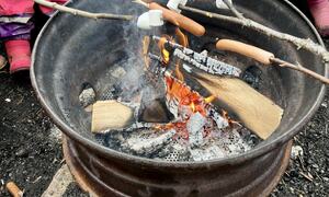 Barn steker pølser og marshmallows over et bål, iført fargerikt regntøy og sikkerhetsvester under en utendørsaktivitet