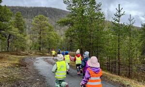 En gruppe barn i fargerike sikkerhetsvester løper på en skogssti, omgitt av trær og fjell