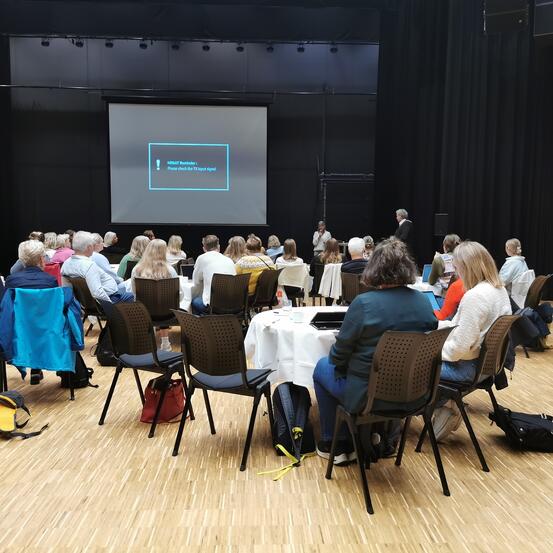 Masse folk sitter i salen rund flere bord og ser på skjermen