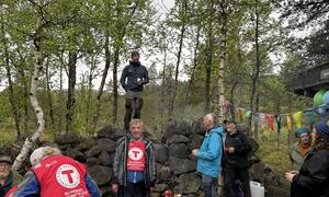 Gruppe samlet rundt utendørs matlagingsområde i skogen, med en person som står på en steinmur
