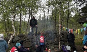 Gruppe samlet rundt bål i skoglysning. Fargerike flagg hengende mellom trærne. Folk lager mat og sosialiserer blant steiner og bjørketrær