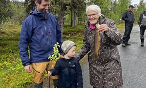 Tre generasjoner på naturtur: mann i blå jakke, barn som holder en plante, eldre kvinne med fisk, skogbakgrunn