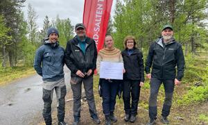 Tre menn og to kvinner står i skogen, flagg bakerst, en kvinne holder gavesjekk, alle smiler