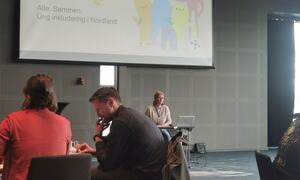 En mann og en kvinne sitter ved bordet, andre kvinne står på scene