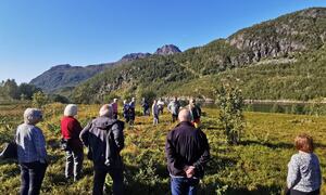 Folk står ute i naturen og hører på en mann, fjell i bakgrunn