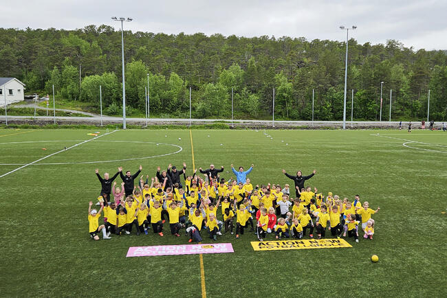 Masse barn og flere voksne i sportsklær på footballbane står samles med hendene opp