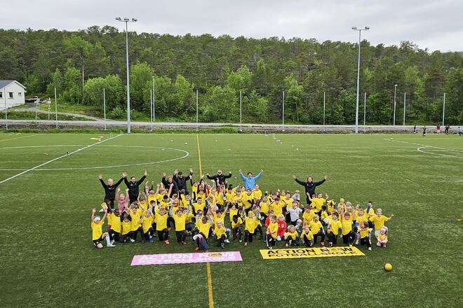 Masse barn og flere voksne i sportsklær på footballbane står samles med hendene opp