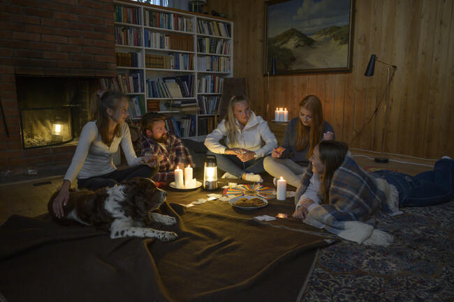 Strømbrudd. Fem unge mennesker sitter i stua foran peisen med pledd rundt seg og spiller spill.