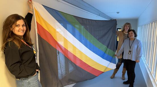 Silje, Janne og Stine holder frem funksjonshemmedes flagg som består av en sort bakgrunn med fem diagonale striper.