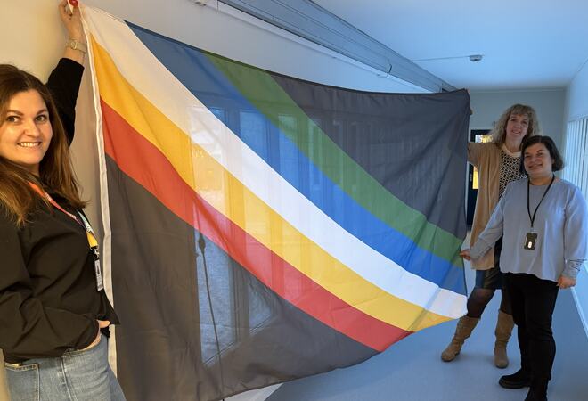 Silje, Janne og Stine holder frem funksjonshemmedes flagg som består av en sort bakgrunn med fem diagonale striper.